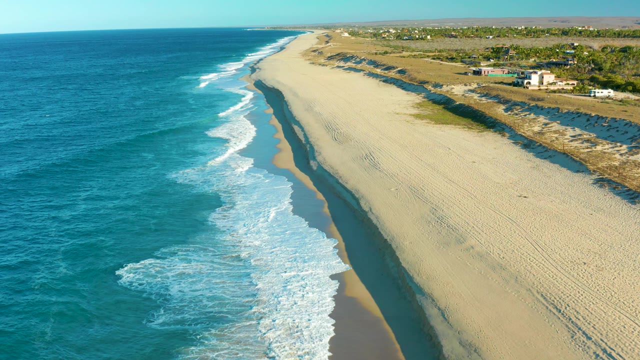 Aerial view in Todos Santos with more whitewater gathered near the lower left, a long strip of wet sand tracing the surf, broad beach to the right, and no visible people.