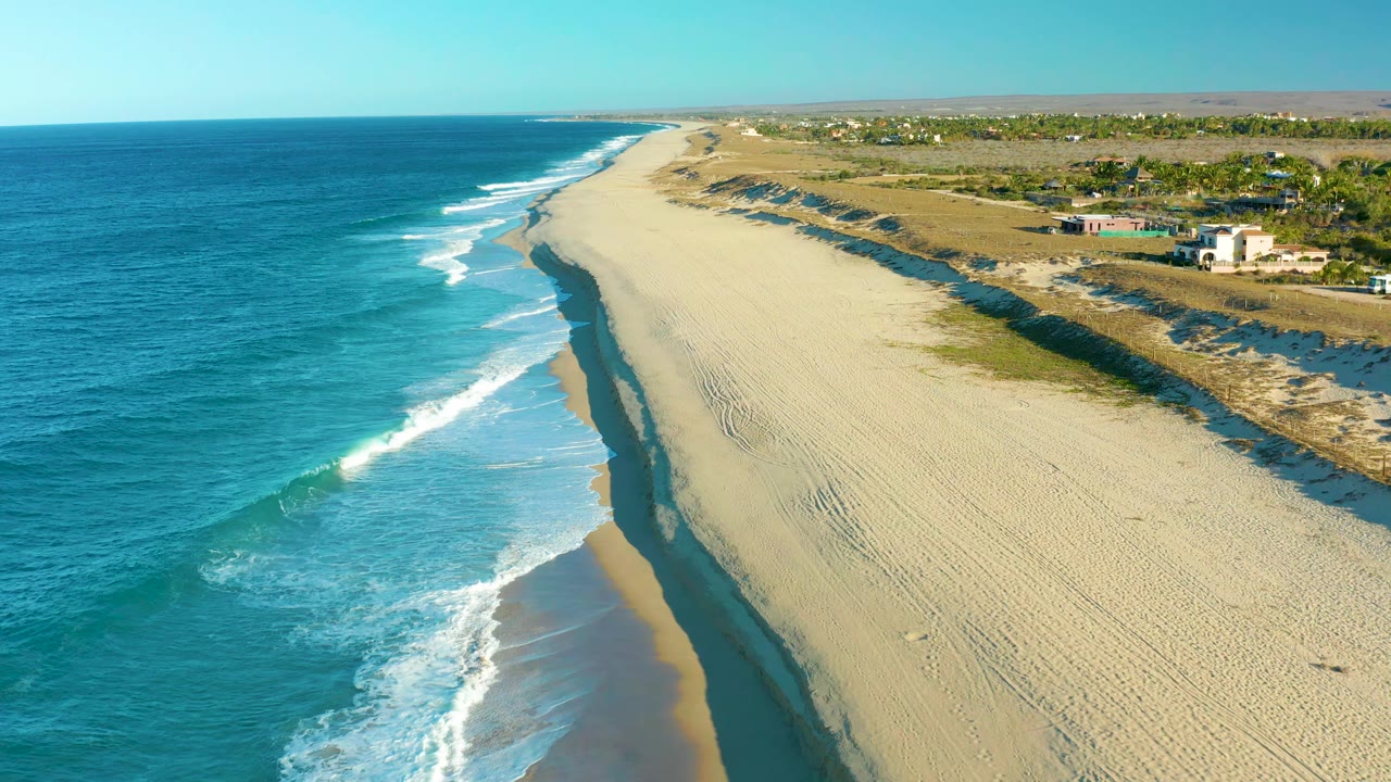 Aerial frame in Todos Santos showing a fresh band of white foam moving onto the beach, turquoise water along the left edge, pale sand through the center, dunes and small inland buildings, and no visible people.