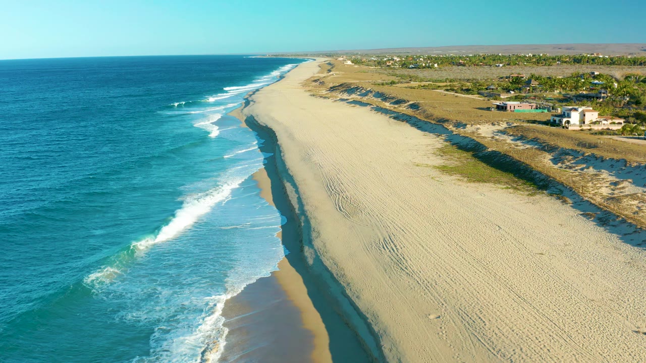 Todos Santos coastline from above, with the wave break slightly farther up the shore, deep blue water offshore, a long uninterrupted sand strip, low dune shadows, scattered vegetation, distant structures, and no visible people.