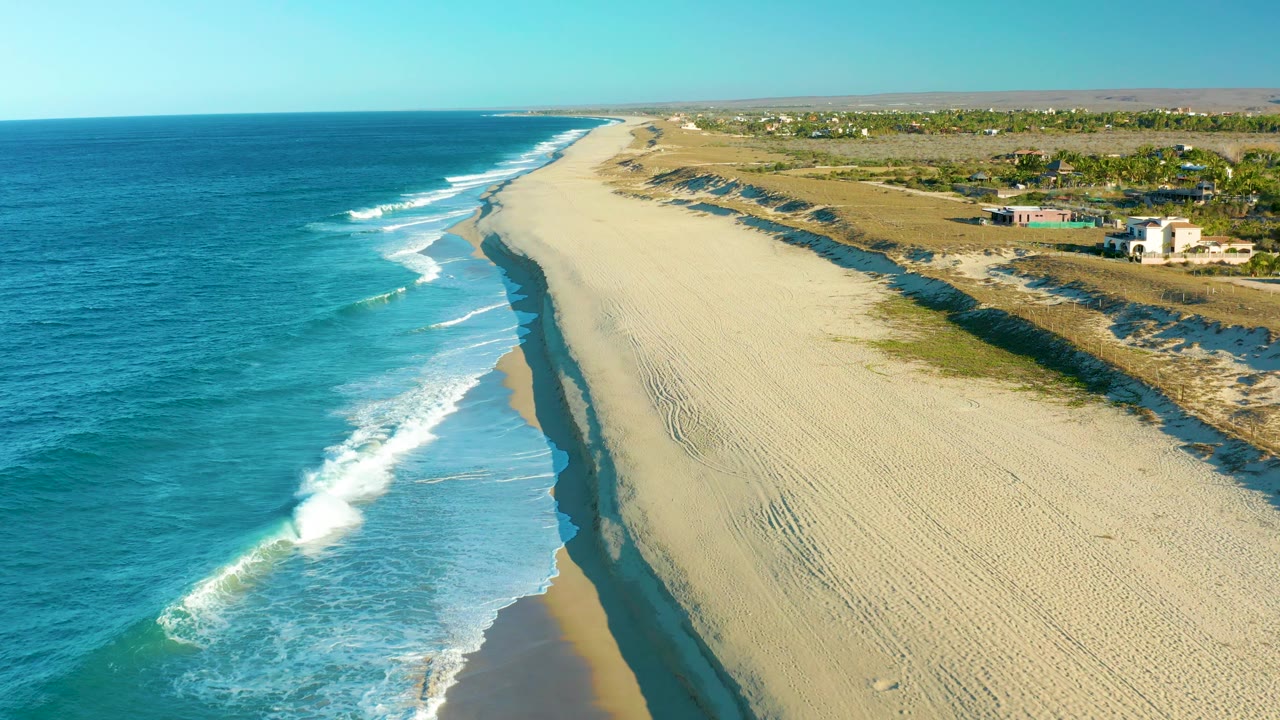 Mid-clip aerial view over Todos Santos beach, with foamy water advancing along the left shoreline, pale raked-looking sand in the center, a raised dune edge to the right, sparse inland growth, small buildings, and no visible people.
