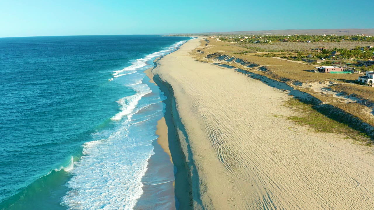 A high aerial view over Todos Santos, Baja California Sur shows blue ocean at left, white surf reaching a wide sandy beach, dunes on the right, and no visible people.