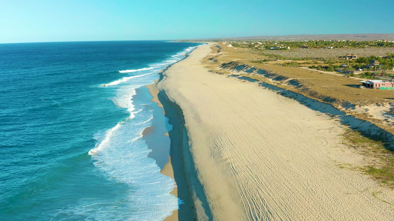 Aerial frame over Todos Santos showing a longer run of foamy surf along the left edge of the pale beach, with dune shadows and scrubland continuing inland and no visible people.