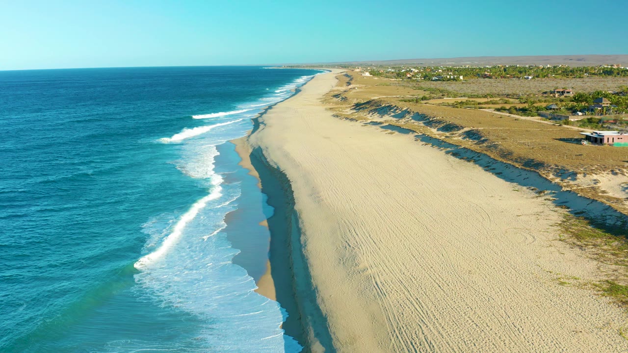 The Todos Santos shoreline remains angled through frame, with turquoise water and a bright breaking wave on the left, empty sand across the center, dune ridges to the right, and no visible people.