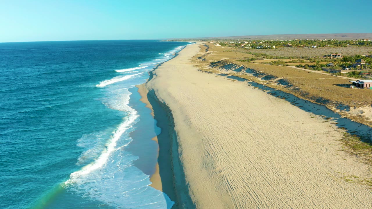 Mid-clip aerial view in Todos Santos with the surf line curving closer to the lower left, the beach stretching toward the horizon, low dunes on the inland side, and no visible people.