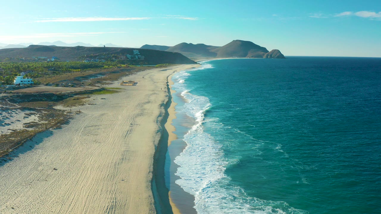 A high aerial frame over Todos Santos, Baja California Sur shows a long sandy beach, turquoise ocean surf, inland scrub, distant hills, and no visible people.