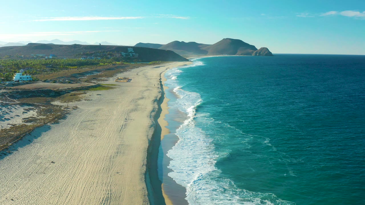 The next aerial frame keeps the Todos Santos beach centered, with foamy surf advancing along the right edge, pale sand below, green vegetation inland, and no visible people.