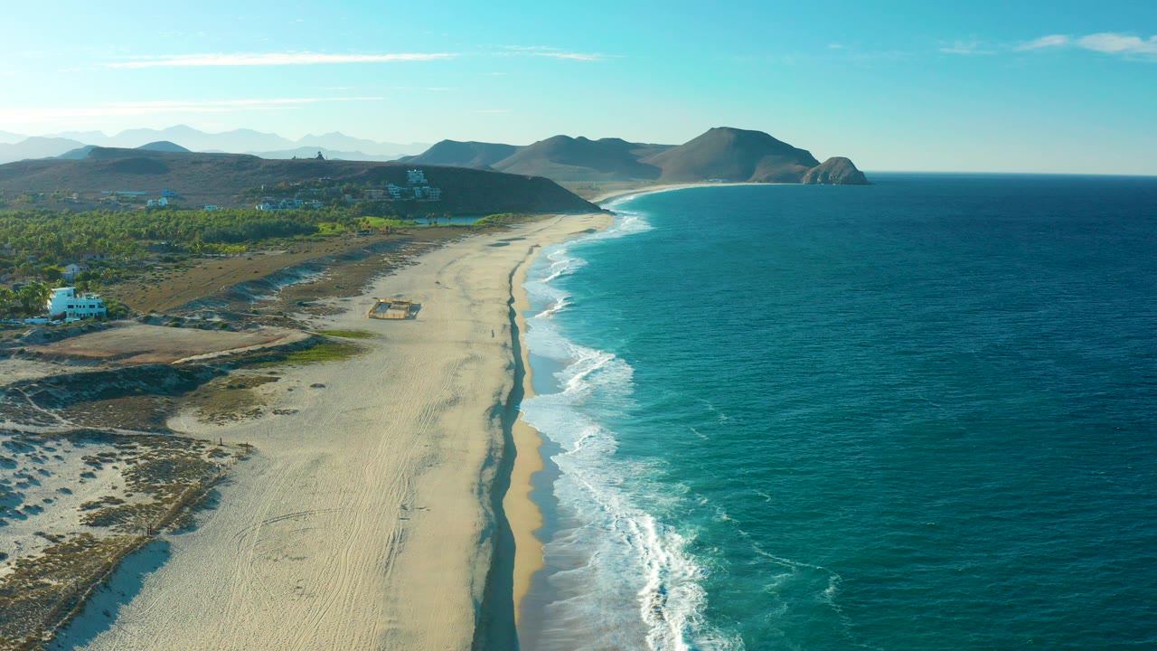 Aerial view over Todos Santos, Baja California Sur, with a broad sandy beach running beside blue surf, inland vegetation and buildings to the left, distant hills on the horizon, and no visible people.