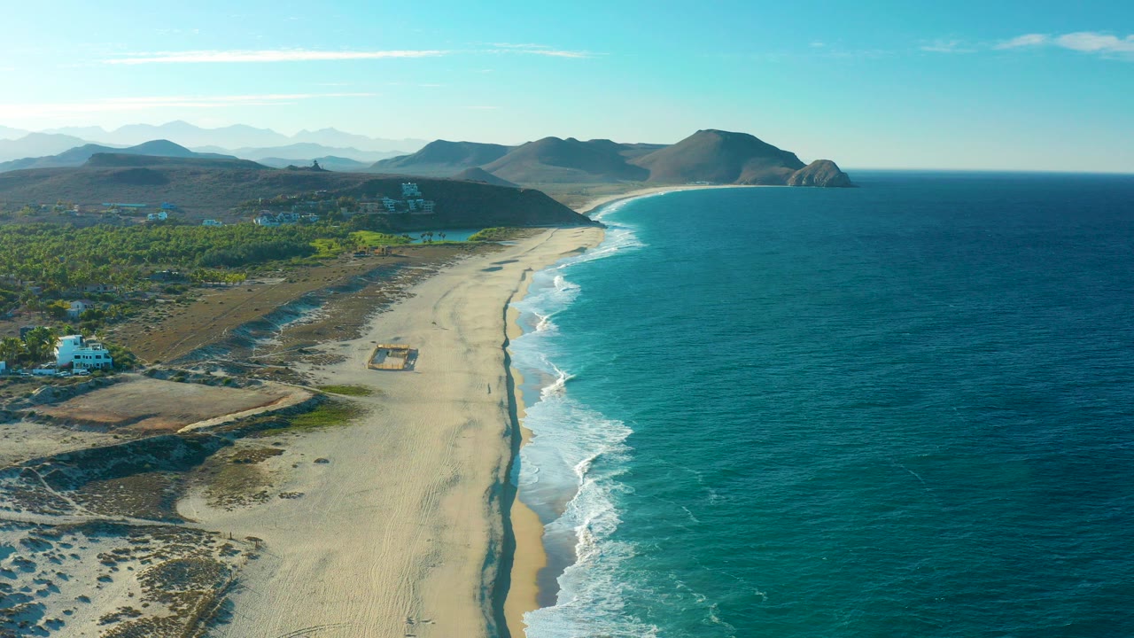 Aerial frame from Todos Santos, Baja California Sur, Mexico, showing a long sandy beach curving beside blue ocean, inland greenery, low hills, clear sky, and no visible people.