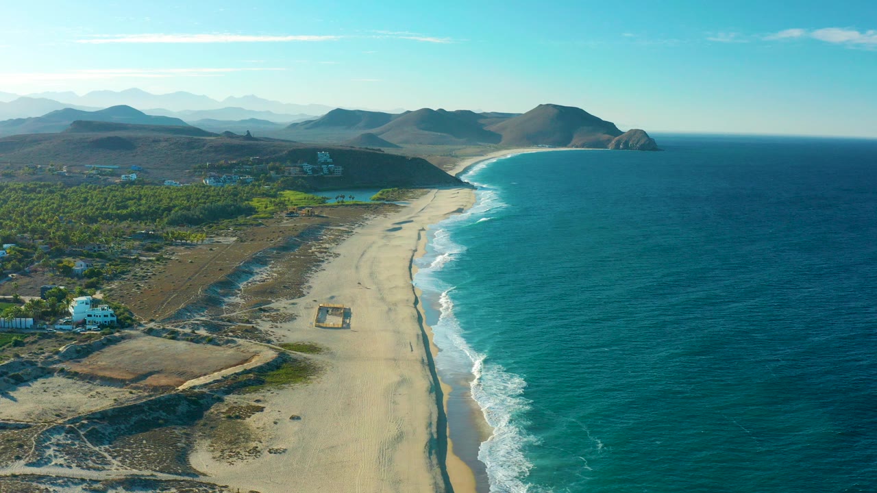 Aerial view over Todos Santos, Baja California Sur with a sandy beach curving beside turquoise ocean, green coastal land on the left, distant hills on the horizon, and no visible people.