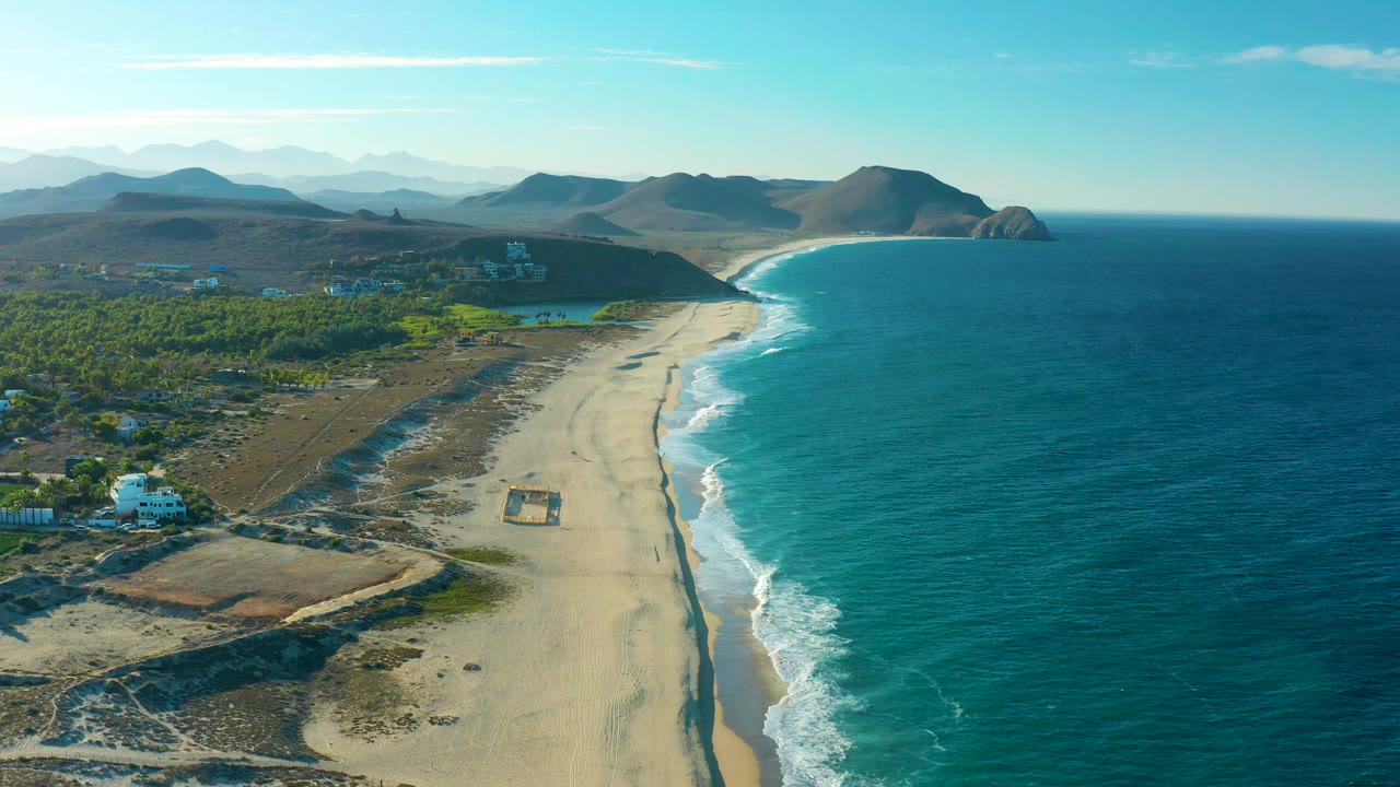 Wide drone view of surf running along a long beach with inland greenery and hills.