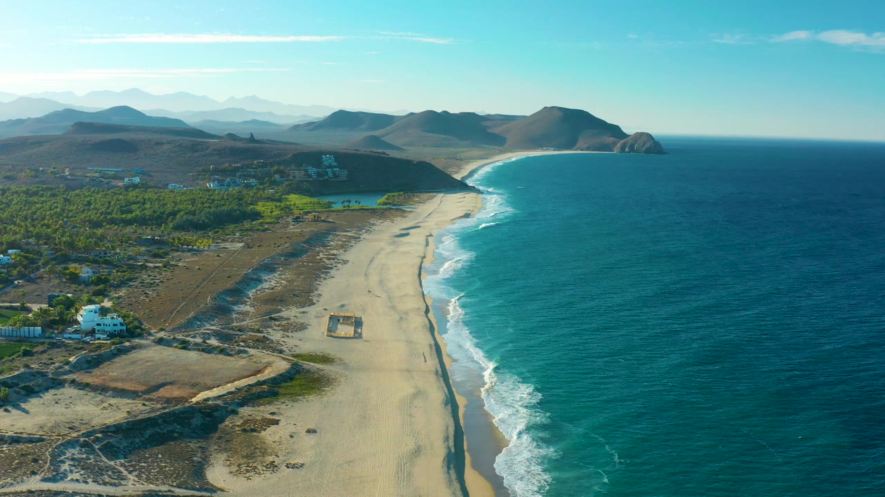 Aerial frame of Todos Santos with deep blue-green ocean on the right, pale sand and darker wet tide marks through the center, inland greenery to the left, and no visible people.