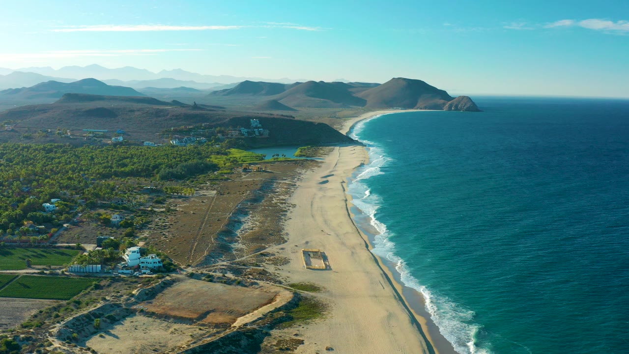 A high aerial view over Todos Santos shows a sandy beach running beside blue ocean, green fields and scattered buildings inland, rolling hills in the distance, and no visible people.