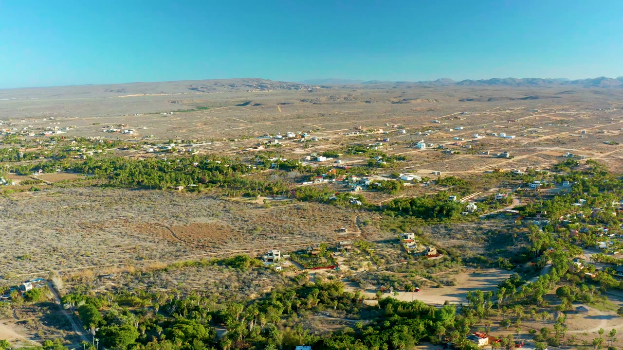 Opening aerial frame over Todos Santos, Baja California Sur, showing scrubby desert foreground, scattered settlement buildings, green tree bands, distant mountains, clear sky, and no visible people.