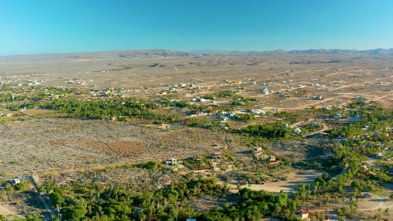 Wide daytime aerial over arid settlement with trees, roads, and mountains beyond.