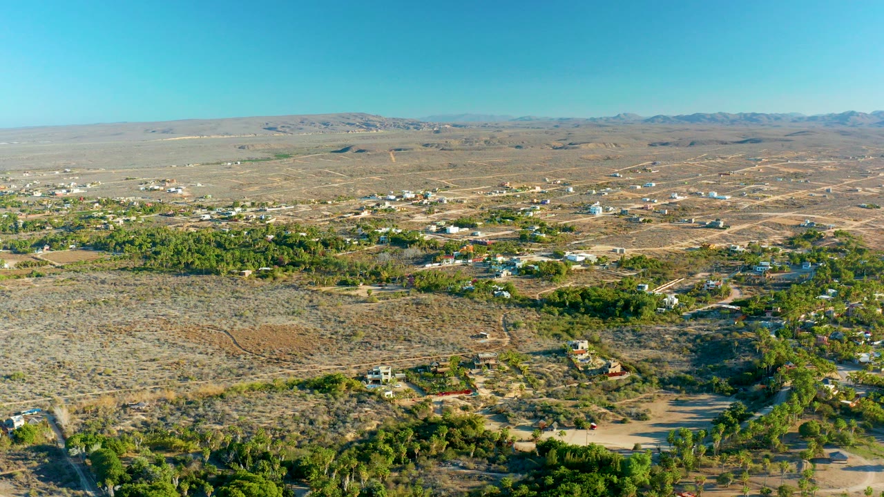 A high aerial frame over Todos Santos, Baja California Sur shows dry terrain, green tree clusters, scattered structures, dirt roads, distant mountains, and no visible people.