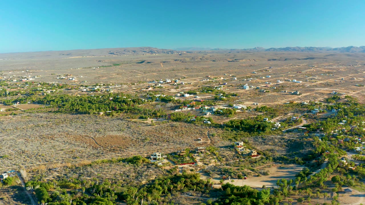 A wide Todos Santos drone view shows arid ground in the foreground, small structures and trees through the middle distance, a mountain line, and no visible people.