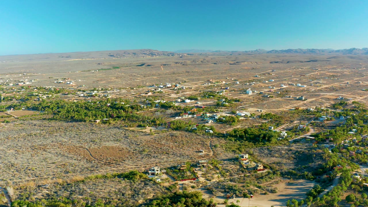 Wide aerial frame over Todos Santos, Baja California Sur, showing dry scrubland, scattered houses, green tree clusters, dirt roads, distant hills, clear blue sky, and no visible people.