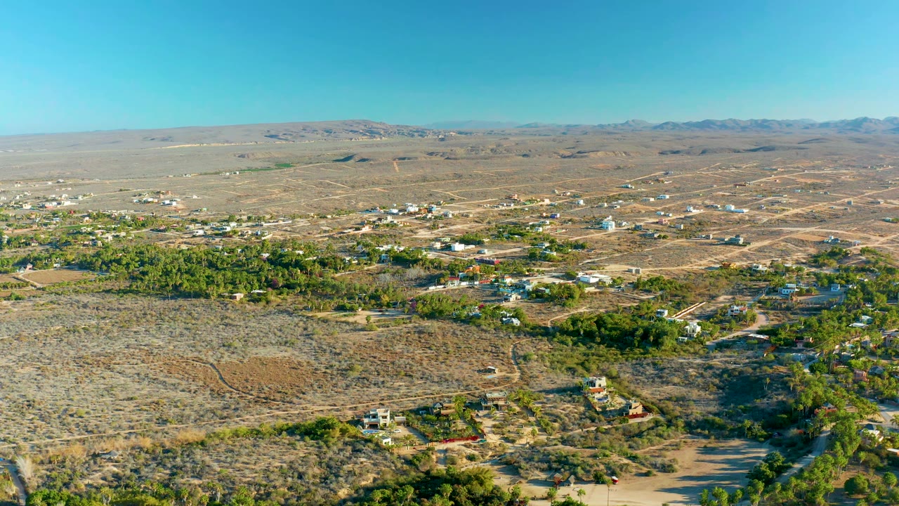 Aerial view in Todos Santos, Baja California Sur, with more of the tree-lined settlement visible below dry open ground, scattered homes, road tracks, low hills, blue sky, and no visible people.