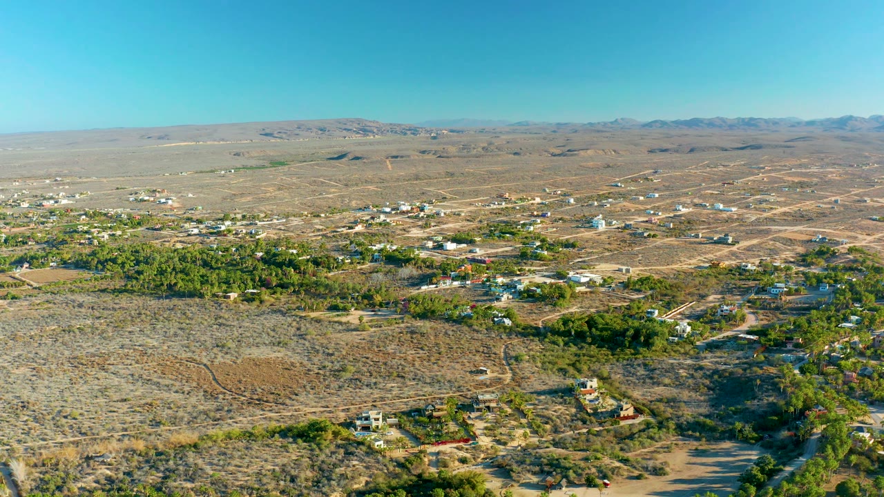High aerial frame over Todos Santos, Baja California Sur, showing a broad band of dry land in front, small homes and trees through the middle, distant hills on the horizon, clear sky, and no visible people.