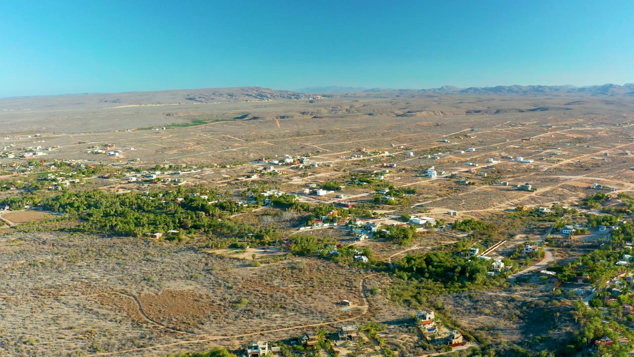 Opening aerial frame over Todos Santos, Baja California Sur, with dry scrub hills in front, scattered homes and trees across the center, distant mountains, clear blue sky, and no visible people.