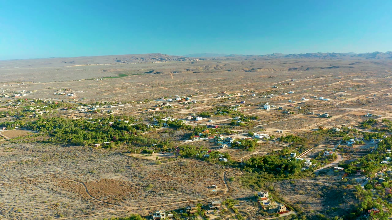 Early aerial frame over Todos Santos showing a broader spread of pale dirt roads, low buildings, green tree clusters, dry open ground, a mountain horizon, and no visible people.