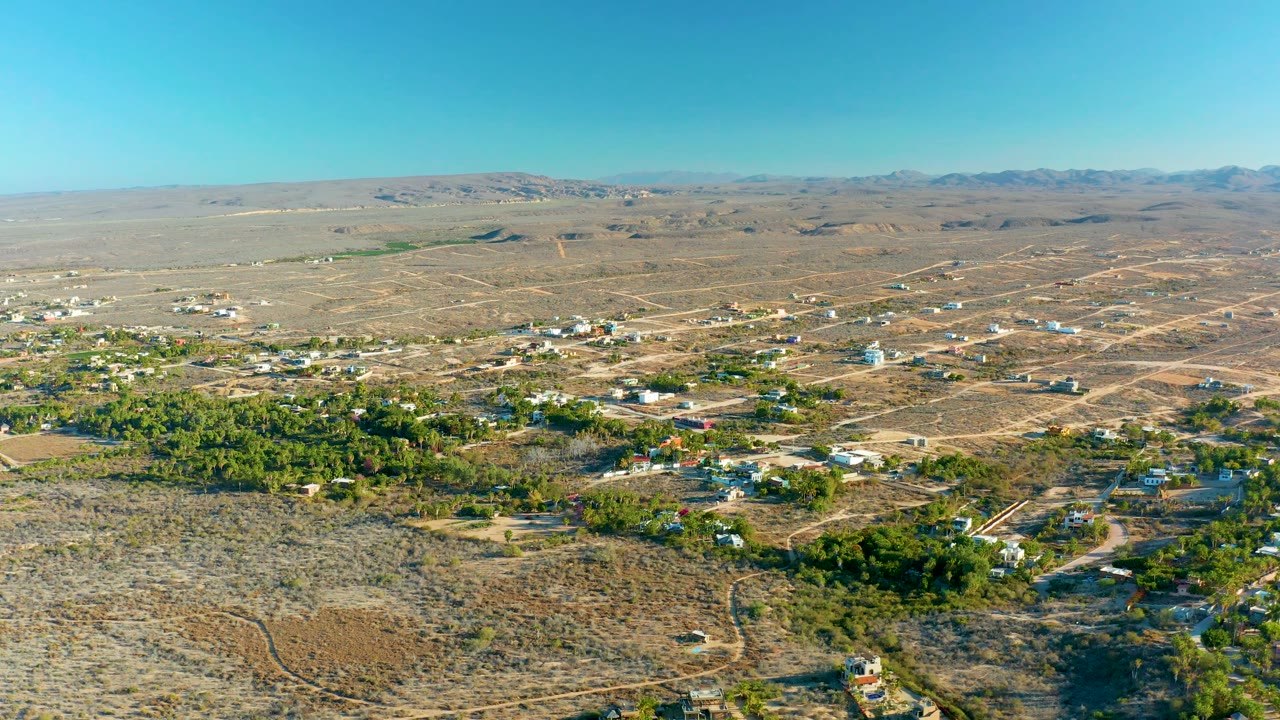 Wide aerial view of dry terrain with homes, dirt tracks, trees, and blue sky.