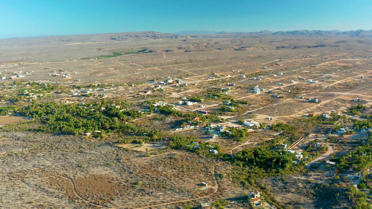 Opening aerial frame over Todos Santos, Baja California Sur, showing tan desert lots, scattered homes, green tree clusters, dirt roads, distant hills, and no visible people.