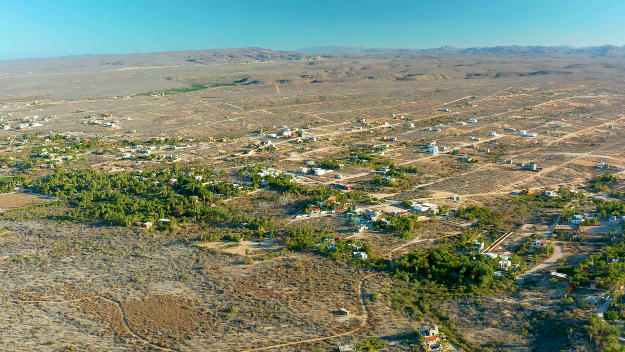Todos Santos aerial view with dusty road lines crossing the desert, sparse homes near green groves, low hills on the horizon, bright daylight, and no visible people.