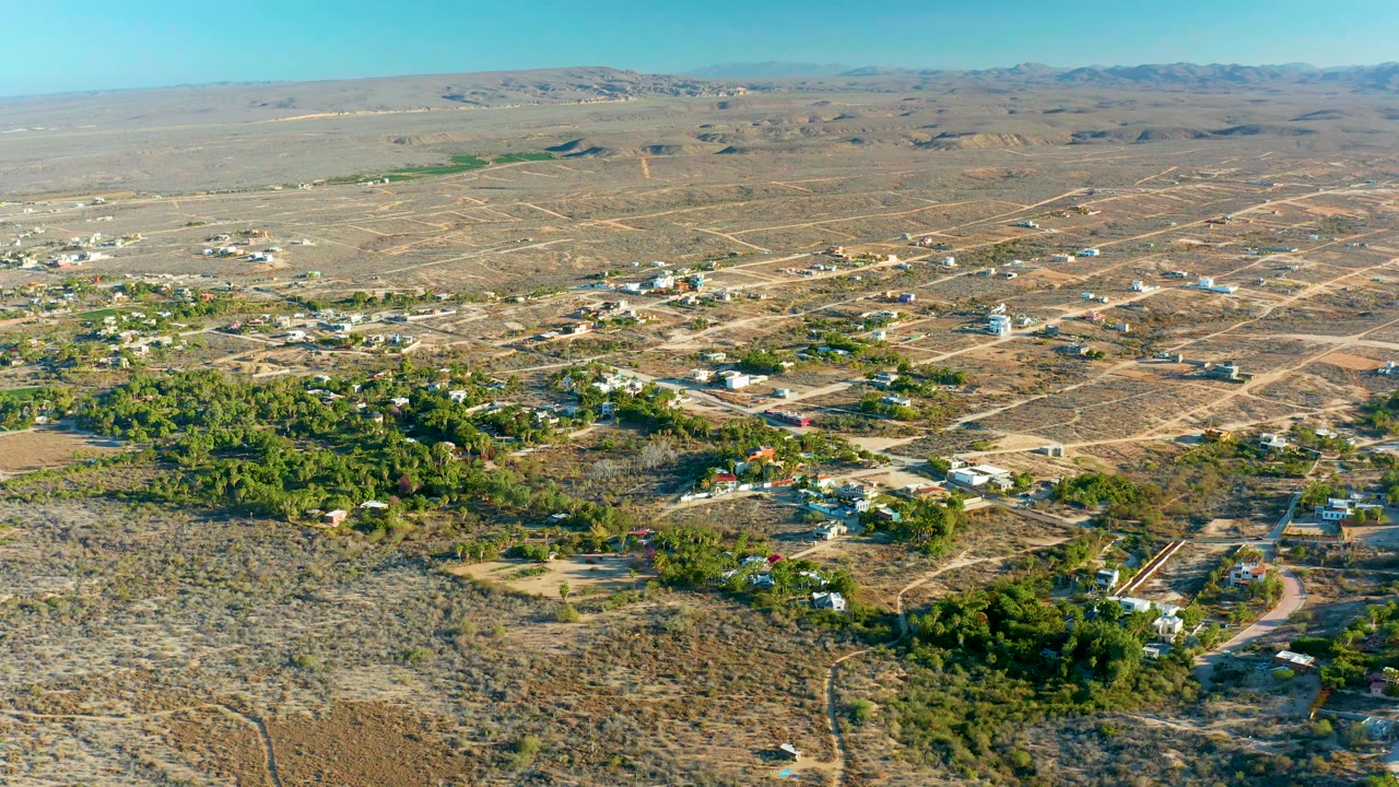 Aerial view in Todos Santos with a green tree pocket near the lower frame, pale roads crossing dry lots, scattered homes beyond it, low hills on the horizon, and no visible people.