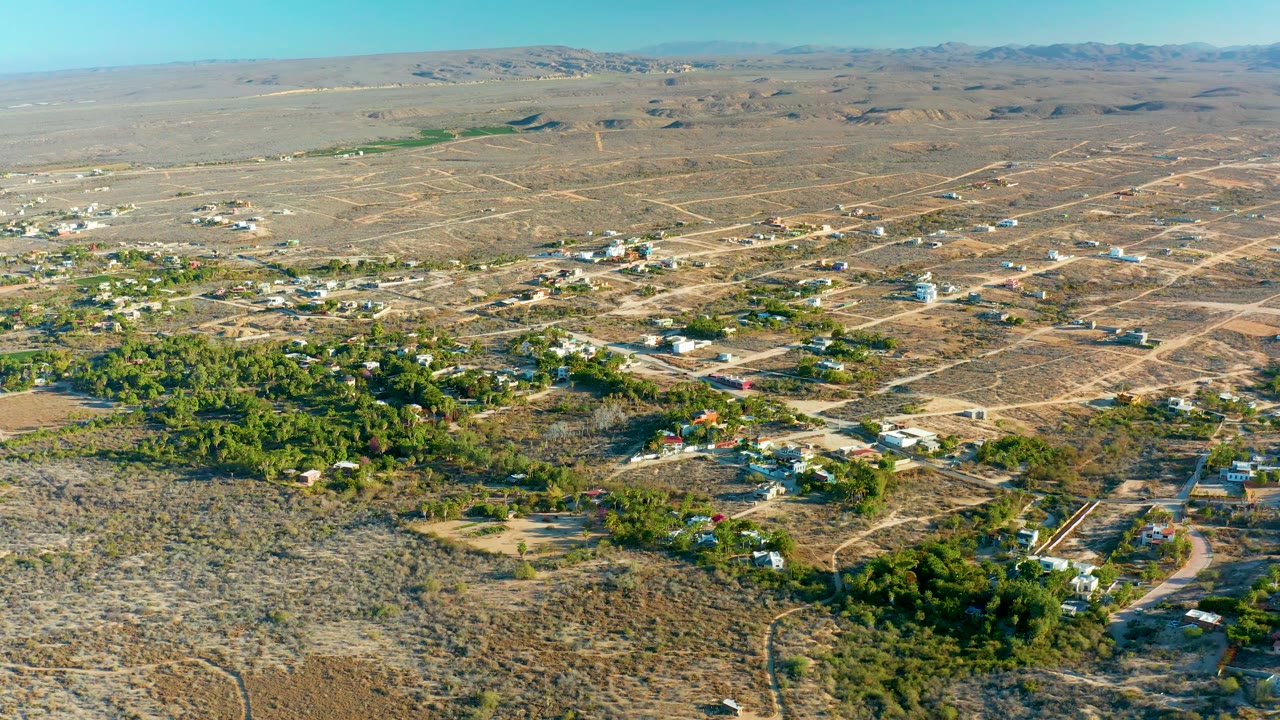 Aerial frame of dry settlement with homes, trees, dirt tracks, and open desert hills.