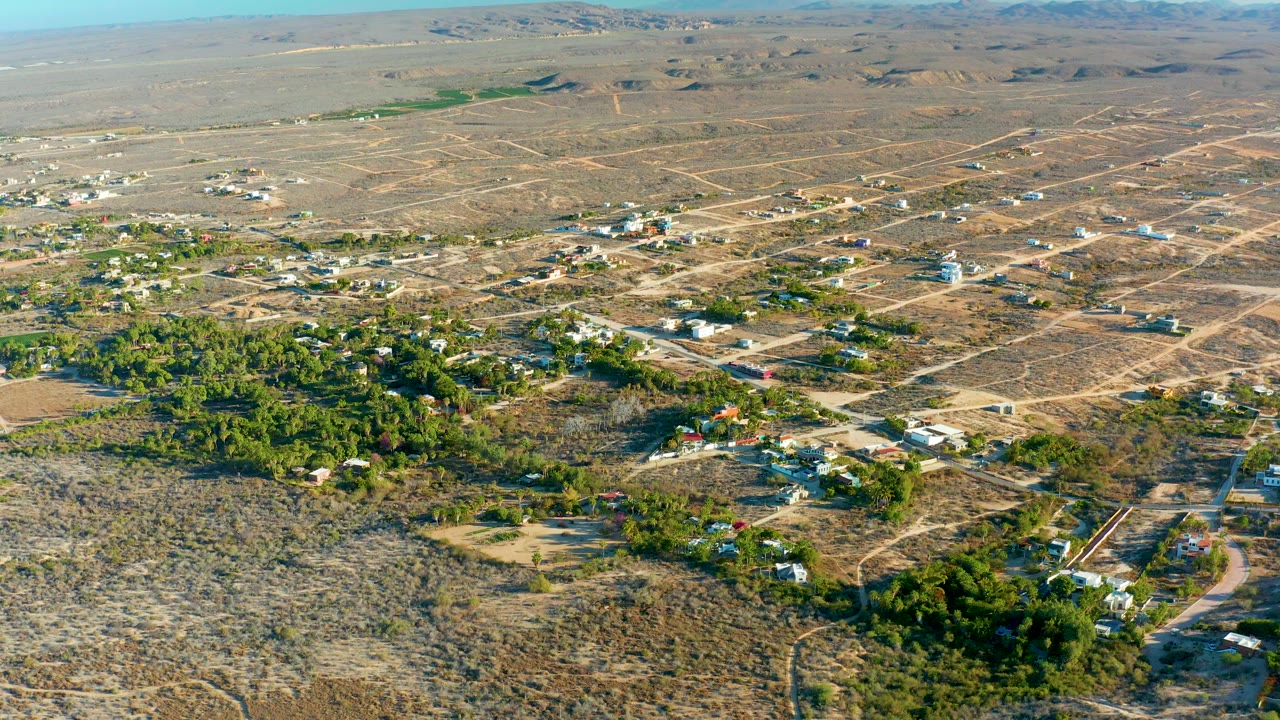 Aerial frame over Todos Santos, Baja California Sur, showing tan desert lots, scattered homes, green tree clusters, distant low hills, and no visible people.