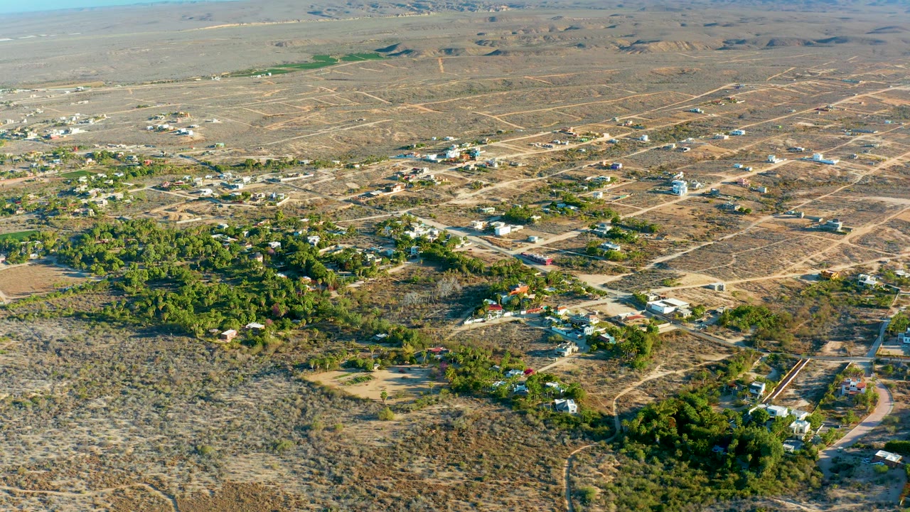 Todos Santos aerial frame with scrubby foreground land, light-colored homes, dirt tracks, dense green vegetation, and no visible people.