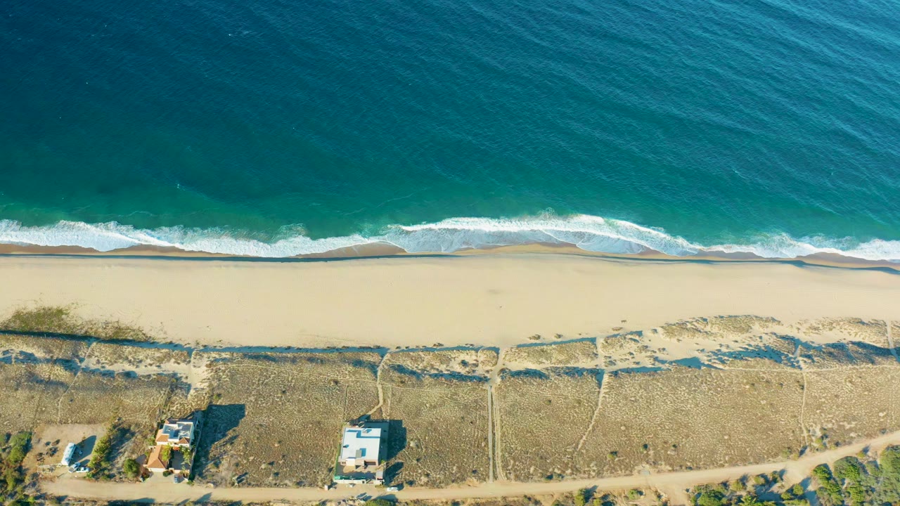 Aerial frame from Todos Santos showing a broader band of turquoise water above the surf, smooth sand through the middle, dune paths, small structures, and no visible people.