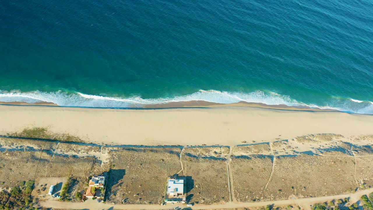 Overhead view in Todos Santos, Baja California Sur, with turquoise water above a white surf line, pale sand, dry inland ground, small houses, and no visible people.