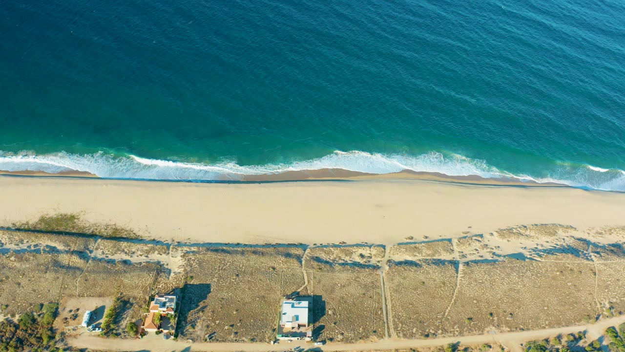 Aerial shoreline with waves washing onto sand and dry ground beyond.