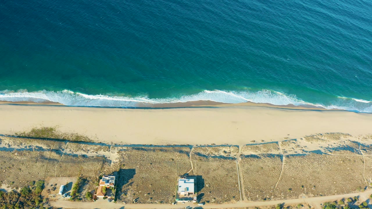 Top-down Todos Santos coastline frame with blue-green ocean texture, a curling white wave, pale beach, dirt access tracks, small houses, and no visible people.