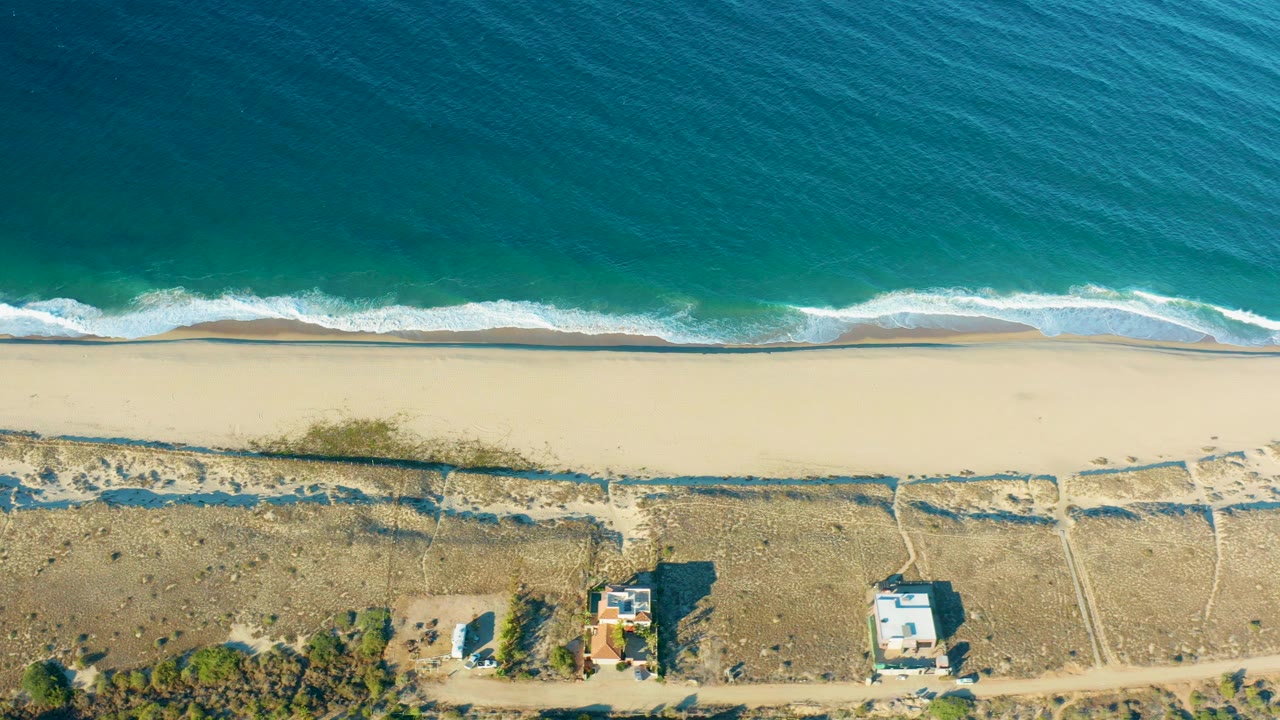 High overhead view of Todos Santos coastline with deep blue water, a thin white surf line, pale sand, dry dunes, dirt paths, small coastal buildings, and no visible people.