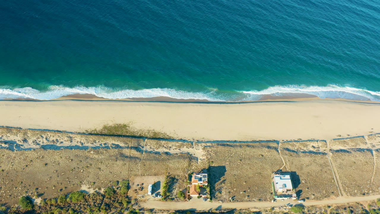Top-down Todos Santos coastal view with a wider band of white breaking surf, smooth sand, scrubby dunes crossed by paths, two small structures, and no visible people.