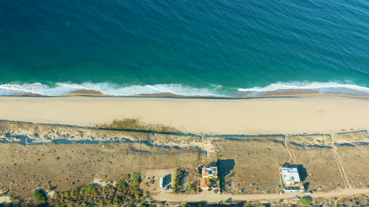 Overhead ocean waves approaching a sandy beach with dunes and scattered buildings.