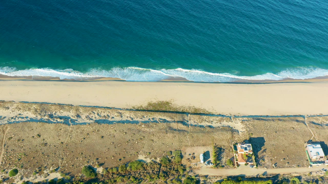 Aerial view of Todos Santos beach as a white wave line reaches the sand, with dry dune terrain, faint paths, shrubs, and a few buildings visible inland, with no visible people.