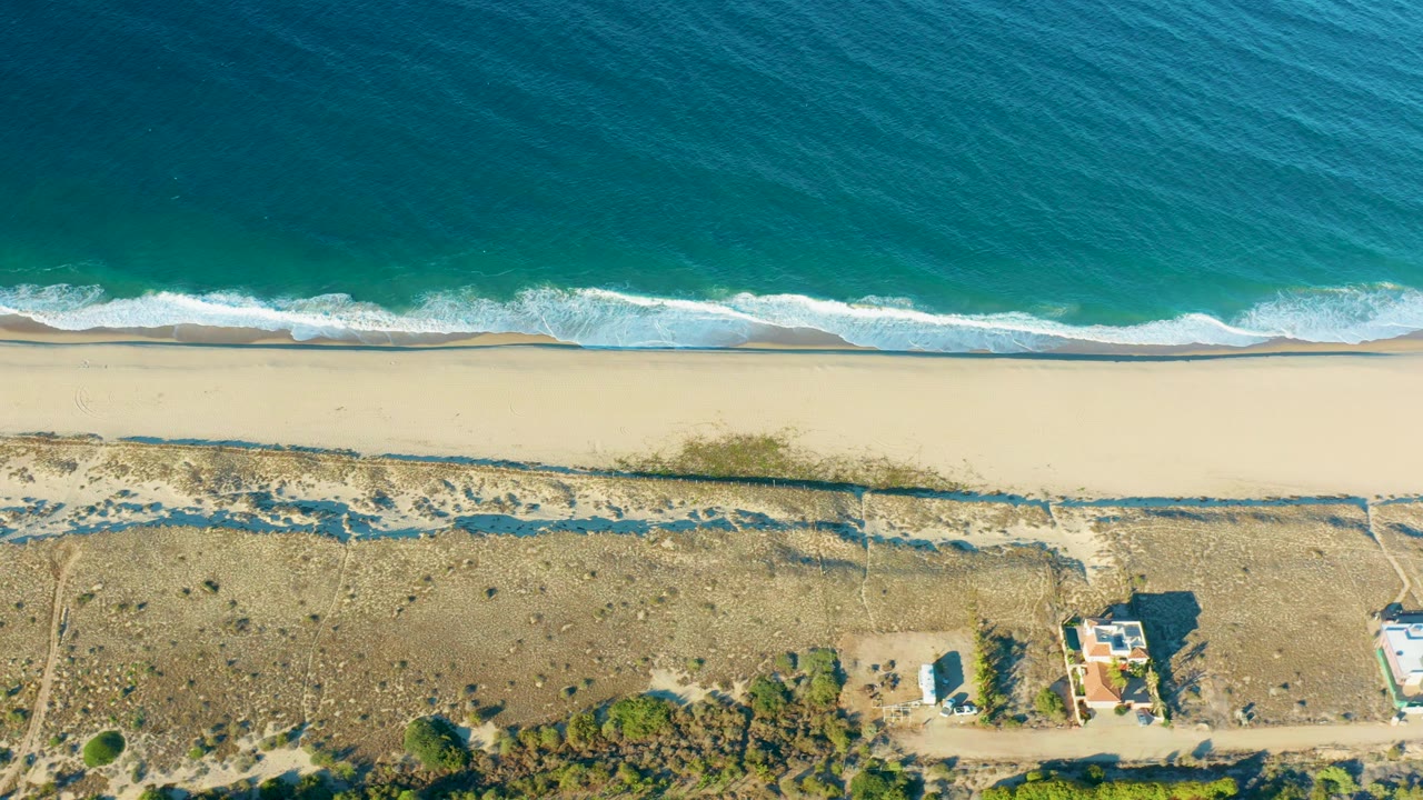 Top-down frame of the Todos Santos coast with textured turquoise water, a continuous foamy surf edge, broad pale beach, dune tracks, low vegetation, and small structures at the bottom, with no visible people.