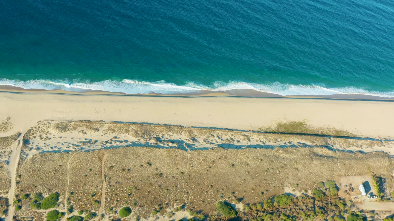 Drone view of Todos Santos coastline with ocean at the top, a continuous surf band, sandy beach, dune paths, scrub patches, a small white structure near the lower right, and no visible people.