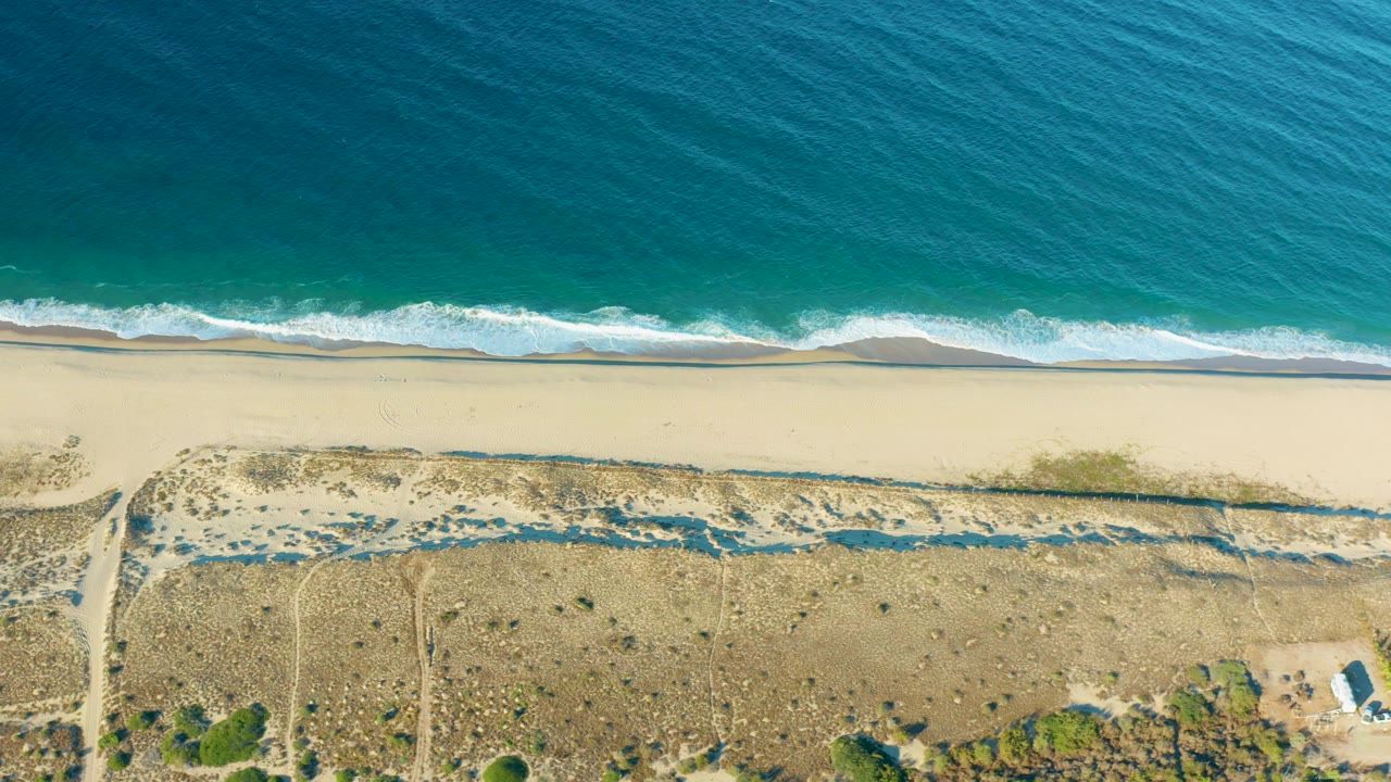 Top-down Todos Santos beach view with turquoise water, a thin white wave line, wide pale sand, dry dunes, access tracks near the frame edges, and no visible people.