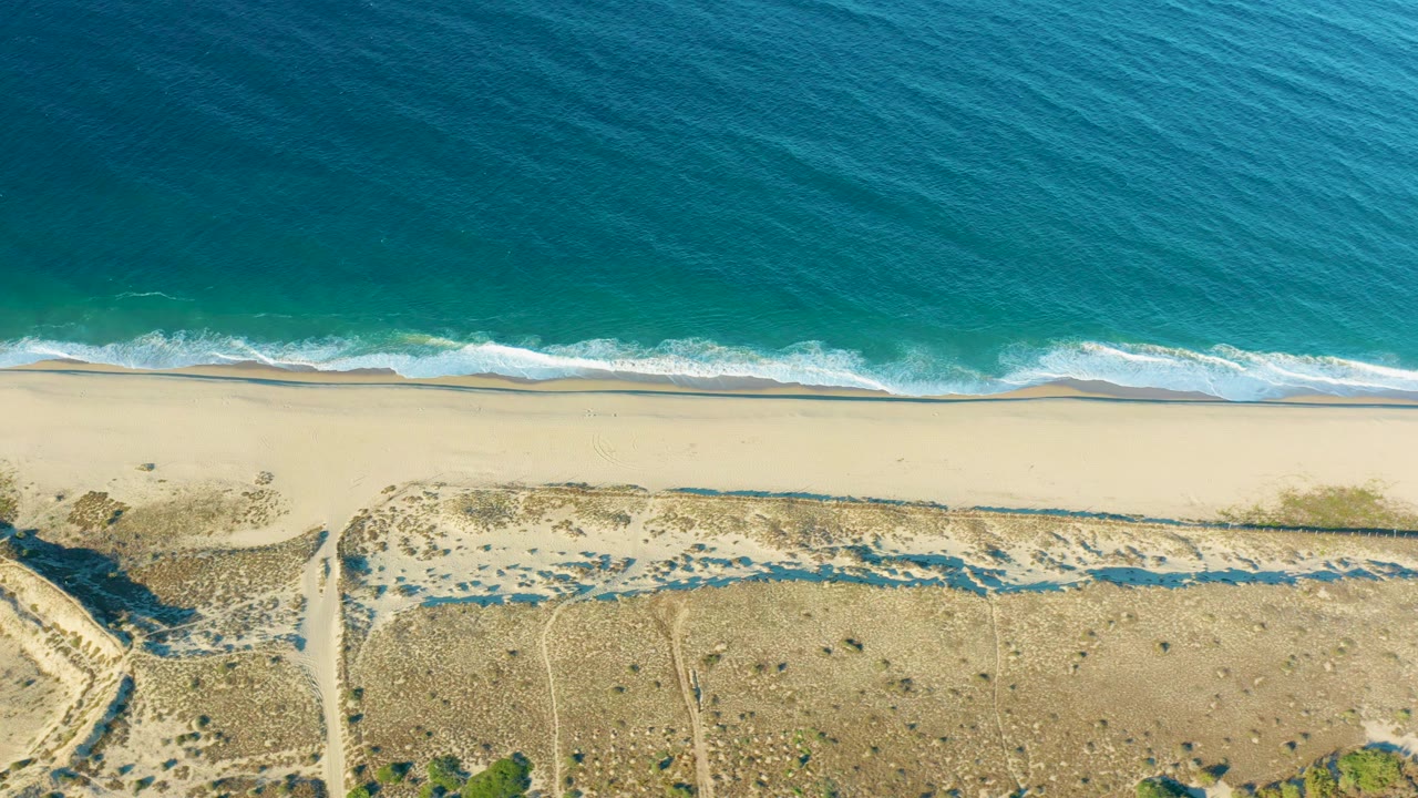 Aerial view of the Todos Santos shoreline where a sandy access path cuts through dry dune terrain below the pale beach and turquoise surf, with no visible people.