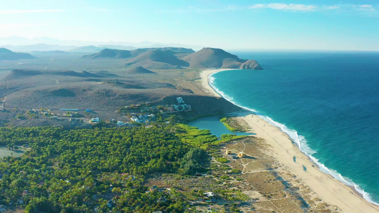 Aerial view of sandy beach, turquoise ocean, trees, lagoon, and coastal hills.
