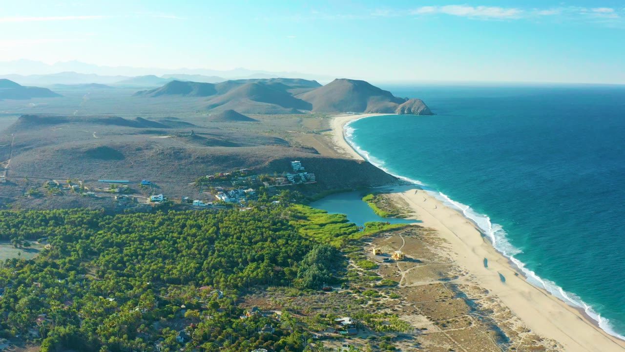 Aerial coastal frame from Todos Santos showing the ocean pushed to the right, the beach and surf line below, green tree cover around the lagoon, dry hills beyond, and no visible people.