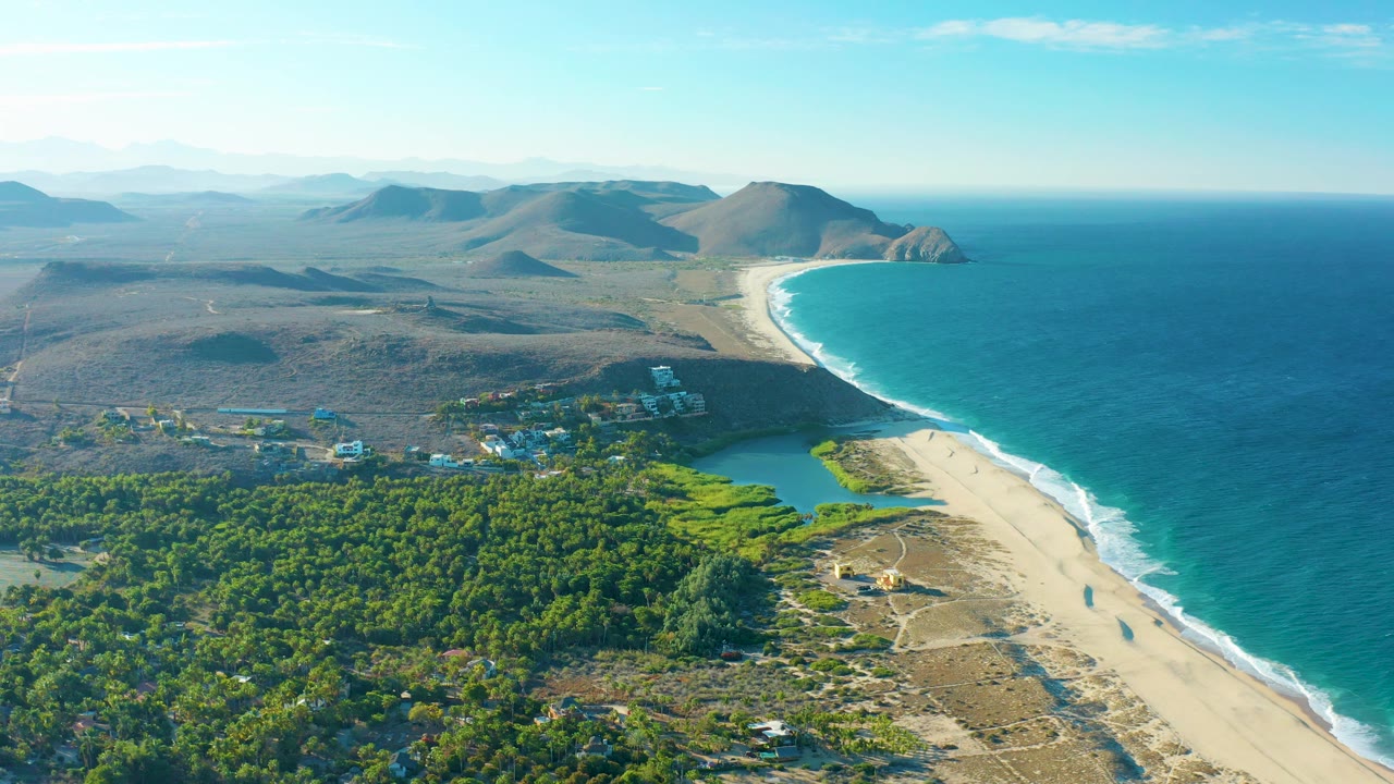 High aerial view over Todos Santos with the beach running diagonally beside blue ocean, the lagoon and trees lower in frame, cloud shadows across the land, and no visible people.