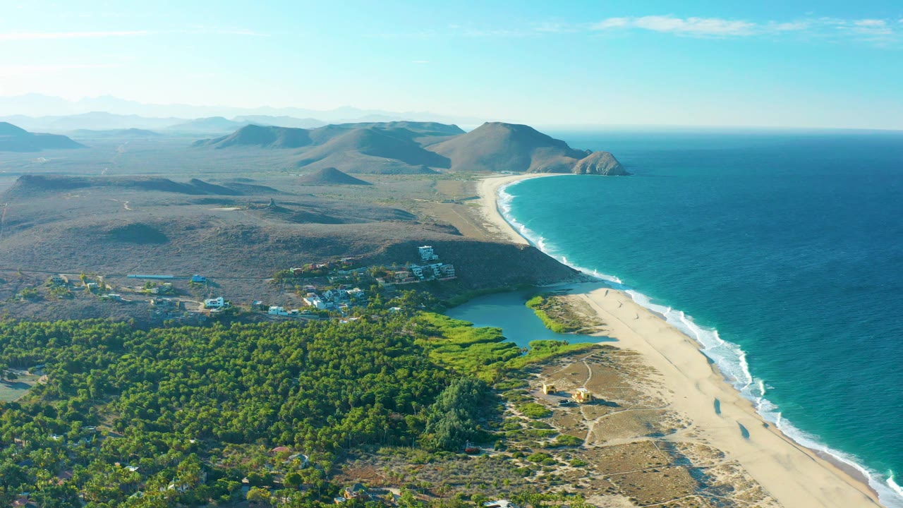 High aerial frame of the Todos Santos coastline showing the beach curve, white waves, green lagoon water, dense coastal vegetation, scattered buildings, distant hills, and no visible people.