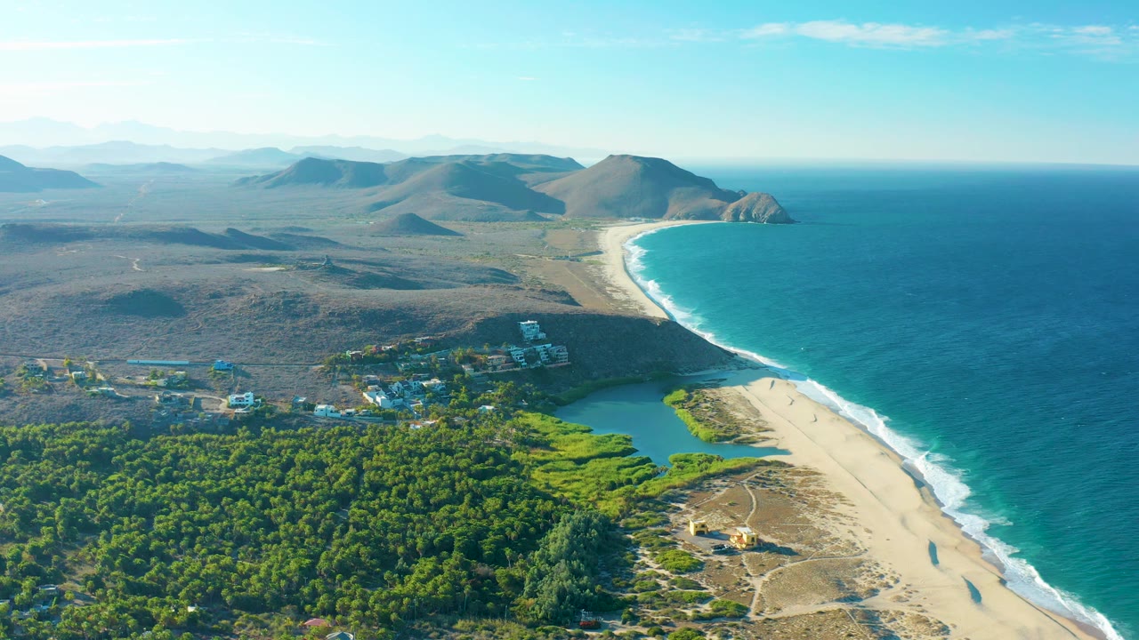 High drone-like view over Todos Santos showing pale sand, breaking waves, a green lagoon, dense coastal vegetation, scattered buildings, distant dry hills, and no visible people.