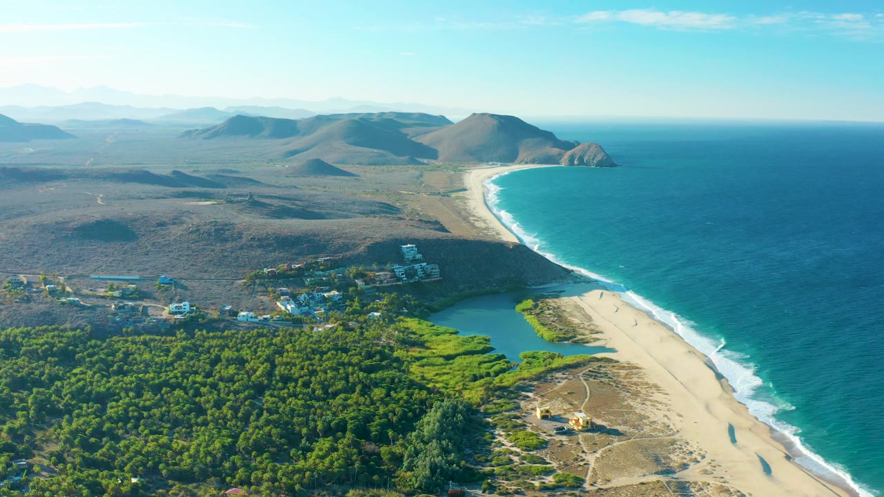 Wide Todos Santos aerial view with the beach running diagonally toward rocky hills, blue ocean to the right, green trees and lagoon below, a small settlement inland, and no visible people.